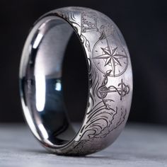 "Celebrate the maritime spirit of your high-seas adventures with this exquisite 8mm wide domed tungsten pirate-themed wedding band. Adorned with iconic nautical symbols including a menacing monsters of the deep, a weathered ship's wheel, a gleaming anchor, and a treasure chest overflowing with doubloons, each meticulously laser etched to create a lasting tribute to your seafaring memories. Our dedication to precision engraving ensures a flawless and sophisticated finish for this ring, featuring wire wheel polishing for a subtle and understated touch. Handcrafted in our humble workshop, using only the finest materials and cutting-edge equipment, each ring is a genuine work of art. Should you desire a custom pirate design that speaks to your unique voyage, do not hesitate to contact us - we' Luxury Round Ring With Compass Design, Vintage Jewelry Men, Pirate Rings, Pirate Wedding Theme, Nautical Symbols, Space Ring, Pirate Wedding, Cool Rings For Men, Ocean Ring