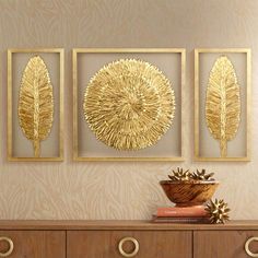 three gold leaf paintings on a wall above a dresser with two vases and books