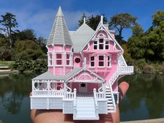 a hand holding a pink and white doll house in front of a body of water