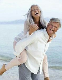 an older man carrying a woman on his back at the beach while they both laugh