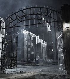 an iron gate with a clock on it in front of a large building at night