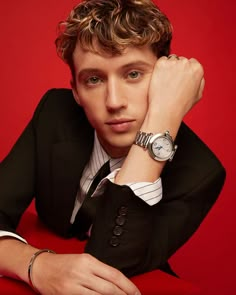 a young man wearing a suit and tie with his hand on his chin, leaning against a red wall