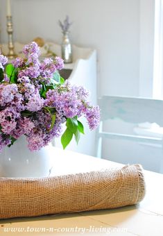 a white vase filled with purple flowers sitting on top of a table next to a piece of burlap