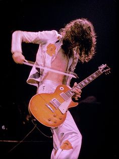 a man with long hair playing an electric guitar while wearing white pants and a button up shirt