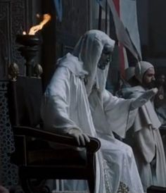 two men dressed in white sitting next to each other on a chair with flags behind them