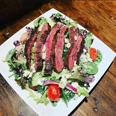 steak salad with lettuce, tomatoes and dressing on a white square plate sitting on a wooden table