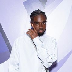 a man with braids standing in front of a purple and white wall holding his hand to his face