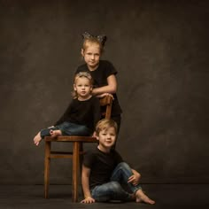 three children sitting on a wooden chair posing for a photo in front of a dark background