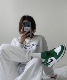 a woman taking a selfie with her cell phone while wearing white sweatpants and green sneakers