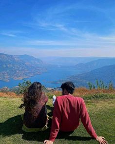 two people are sitting on the grass looking at the mountains and water in the distance