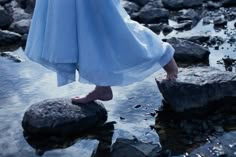 a woman in white dress standing on rocks by water with her feet up to the ground