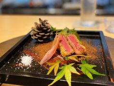 a black plate topped with meat and vegetables on top of a wooden table next to a pine cone