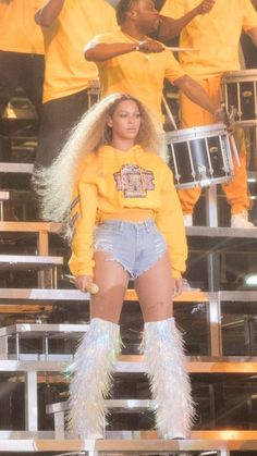 a woman in yellow shirt and shorts standing on stage with other people behind her holding drums