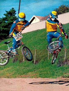 two people on bicycles doing tricks in the air with grass and houses in the background