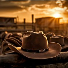 a cowboy hat sitting on top of a wooden rail in front of a fence with the sun setting behind it