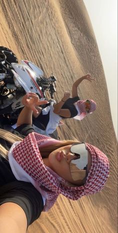 two people riding motorcycles in the desert with sand dunes behind them and one person wearing sunglasses