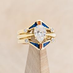 a gold ring with an oval cut diamond and blue sapphire in the center, sitting on top of a wooden stand