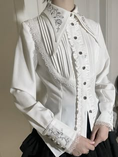 A white button down blouse. The button panel has ruffles down both sides and seven round, silver buttons. The collar is stiff with two layers of fabric and comes to long points. It has silver embroidery of thorns, roses, and crosses. The front of the blouse has pleats in a half oval shape surrounded by lace, descending over the ribs from the shoulders. The sleeves end in cuffs with four buttons on each, silver thorns embroidered, and lace at the ends. Types Of Blouses, Ruffle Outfit, Gothic Shirts, Romantic Blouses, Womens Dress Suits, Blouse Long Sleeve, Collar Designs, Alternative Outfits