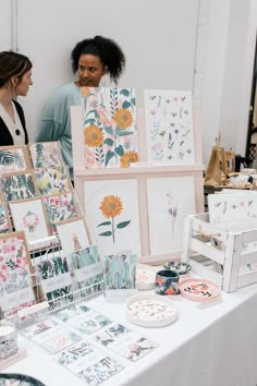 two women standing next to a table full of cards and artwork on it's sides