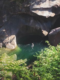 a person swimming in the water near some rocks and trees with green foliage around them