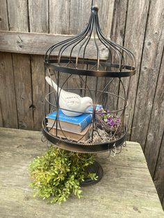 a bird in a cage with books and plants on it sitting on top of a wooden table