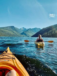 two people in kayaks paddling on the water with mountains in the back ground