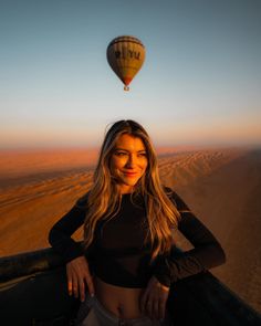 a beautiful woman standing in front of a hot air balloon over a desert landscape at sunset