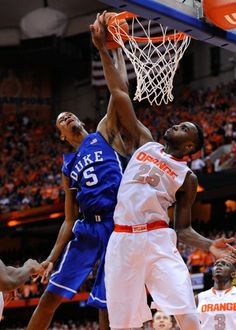 two men playing basketball in front of an orange and white crowd, one is dunking the ball