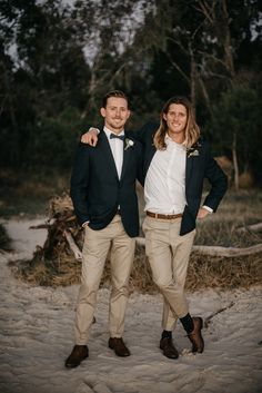 two men standing next to each other in front of some trees and sand with their arms around each other