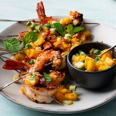 a white plate topped with grilled shrimp next to a bowl of fruit and veggies