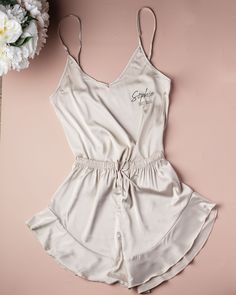 a women's white satin romper with embroidered name on it and flowers in the background