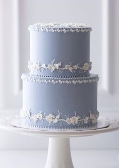 a three tiered blue cake with white flowers on the top and bottom, sitting on a plate