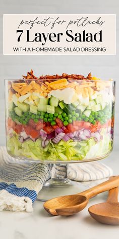 a layered salad in a glass dish with wooden spoons and napkin on the side