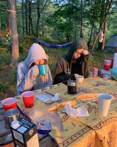 two people sitting at a picnic table with cups in front of them and camping gear on the ground