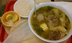 a bowl of soup with meat, corn and rice on a tray next to a spoon