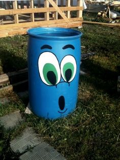 a blue trash can with eyes on it sitting in the grass near a wooden fence