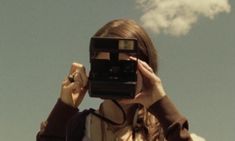 a woman holding up a camera to take a picture with clouds in the sky behind her