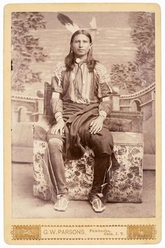 an old photo of a native american man sitting in a chair with feathers on his head