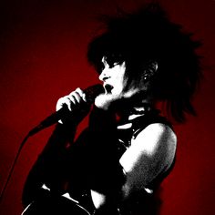 a woman with black hair holding a microphone in front of a red wall and wearing a leather outfit