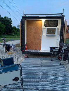 a small trailer is set up in the gravel with two lawn chairs and an open door