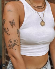 a woman wearing a white tank top with tattoos on her arm and chest, standing in front of a brick wall