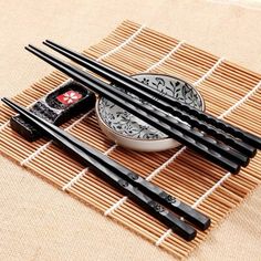 four black chopsticks sitting on top of a bamboo mat next to a bowl