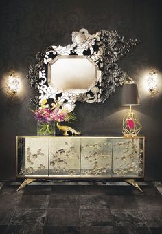 an ornate mirror on the wall above a dresser with flowers in vases and lamps