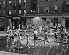 black and white photograph of children playing in water fountain with adults watching from the side