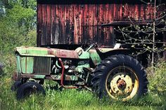 an old tractor sitting in the grass next to a red barn with a rusted roof