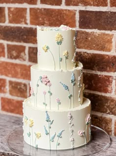 a three tiered cake decorated with flowers on a glass platter next to a brick wall