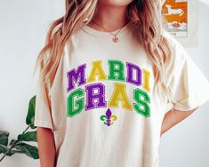 a woman wearing a mardi gras t - shirt standing in front of a plant