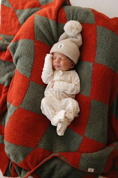 a baby is sleeping on a red and green checkered blanket with a white pom - pom