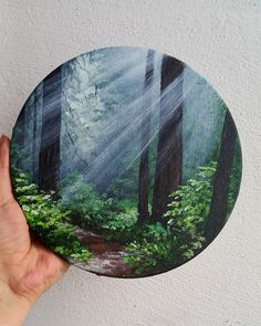 a hand holding up a painting of a forest with sunlight coming through the trees and leaves