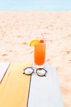 an orange drink sitting on top of a wooden table next to the ocean and beach